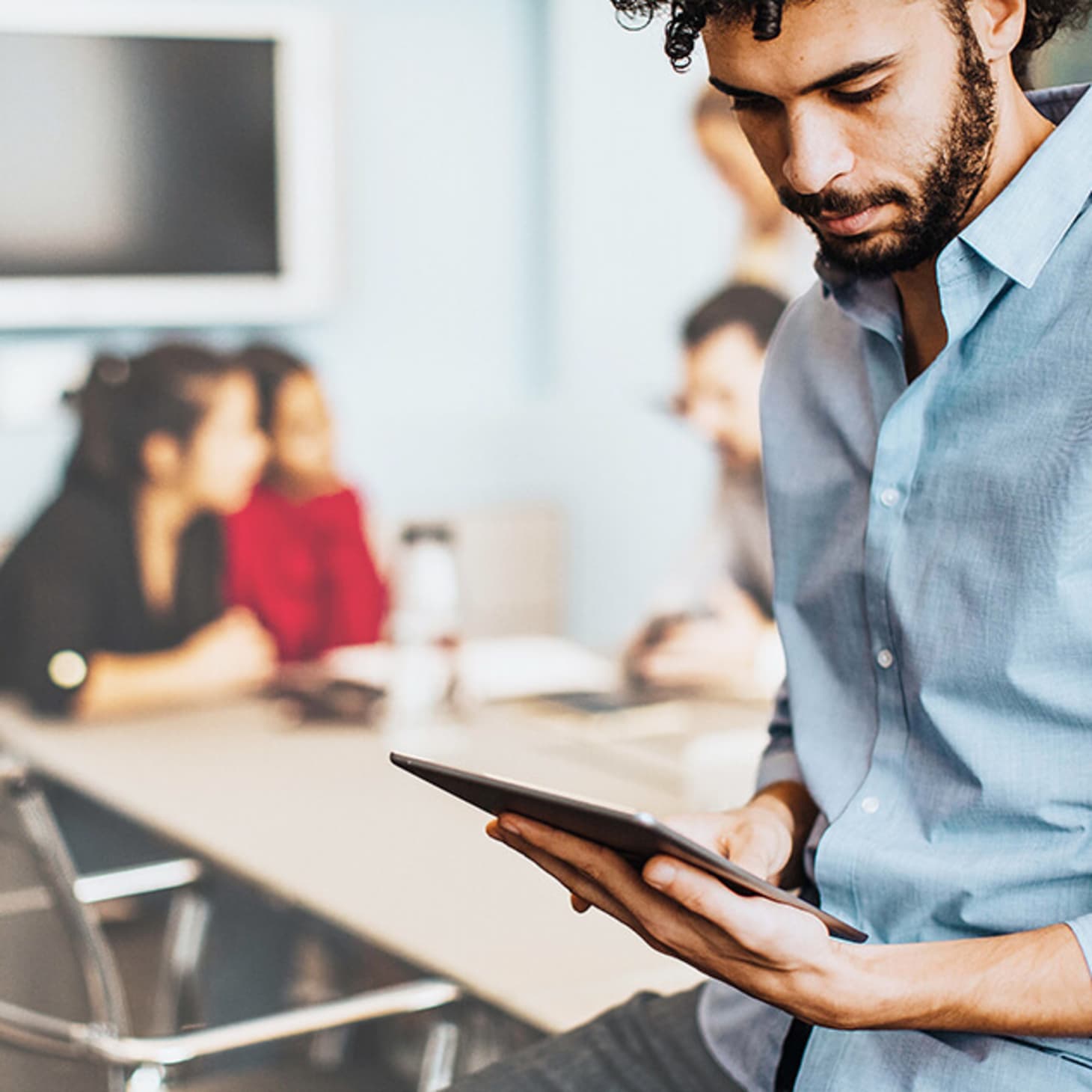 bearded man at office looking at tablet; coworkers in background 