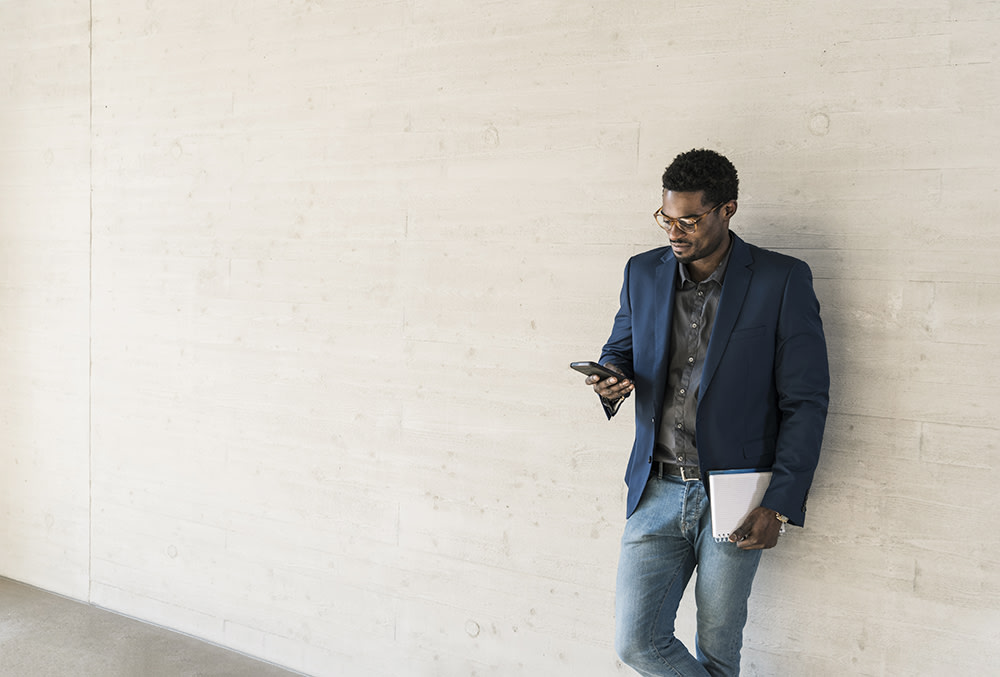 Black man in jeans and suit jacket, leaning against an exterior wall while on his phone